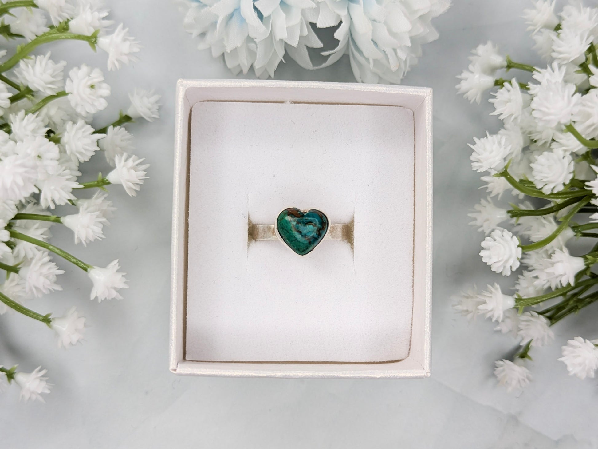 Sterling silver ring with a heart-shaped blue-green chrysocolla gemstone. The ring is in a white jewelry box surrounded by white flowers.
