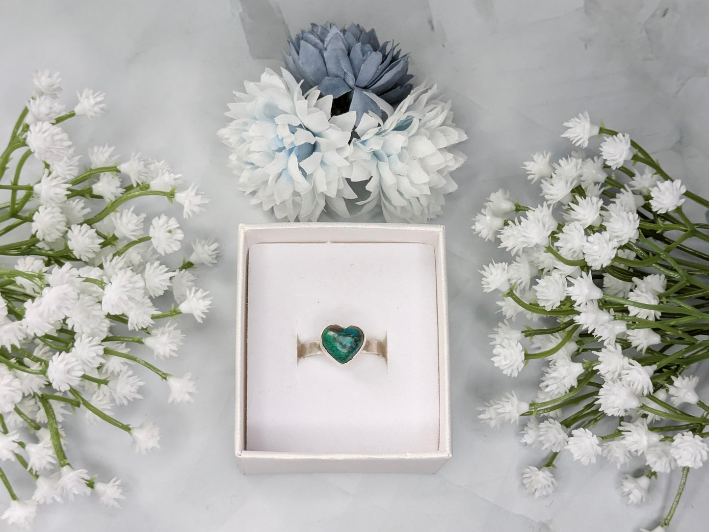 Sterling silver ring with a heart-shaped blue-green chrysocolla gemstone. The ring is in a white jewelry box surrounded by blue and white flowers.