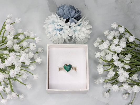Sterling silver ring with a heart-shaped blue-green chrysocolla gemstone. The ring is in a white jewelry box surrounded by blue and white flowers.