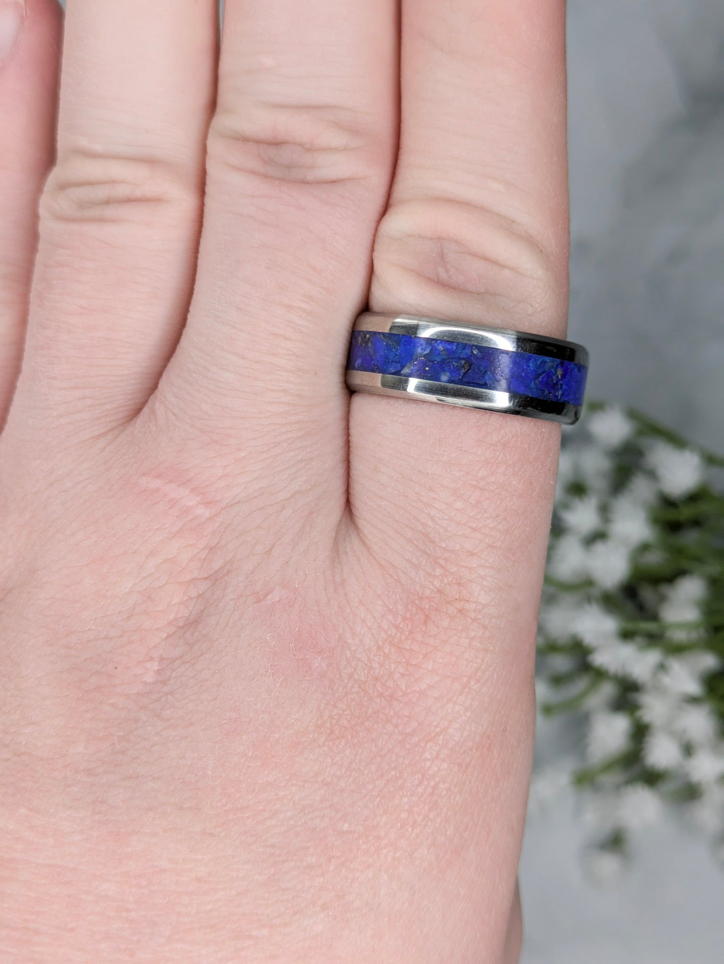 Blue lapis lazuli with flakes of gold pyrite in a stainless steel ring inlay channel. The ring is on a finger with white flowers in the background.