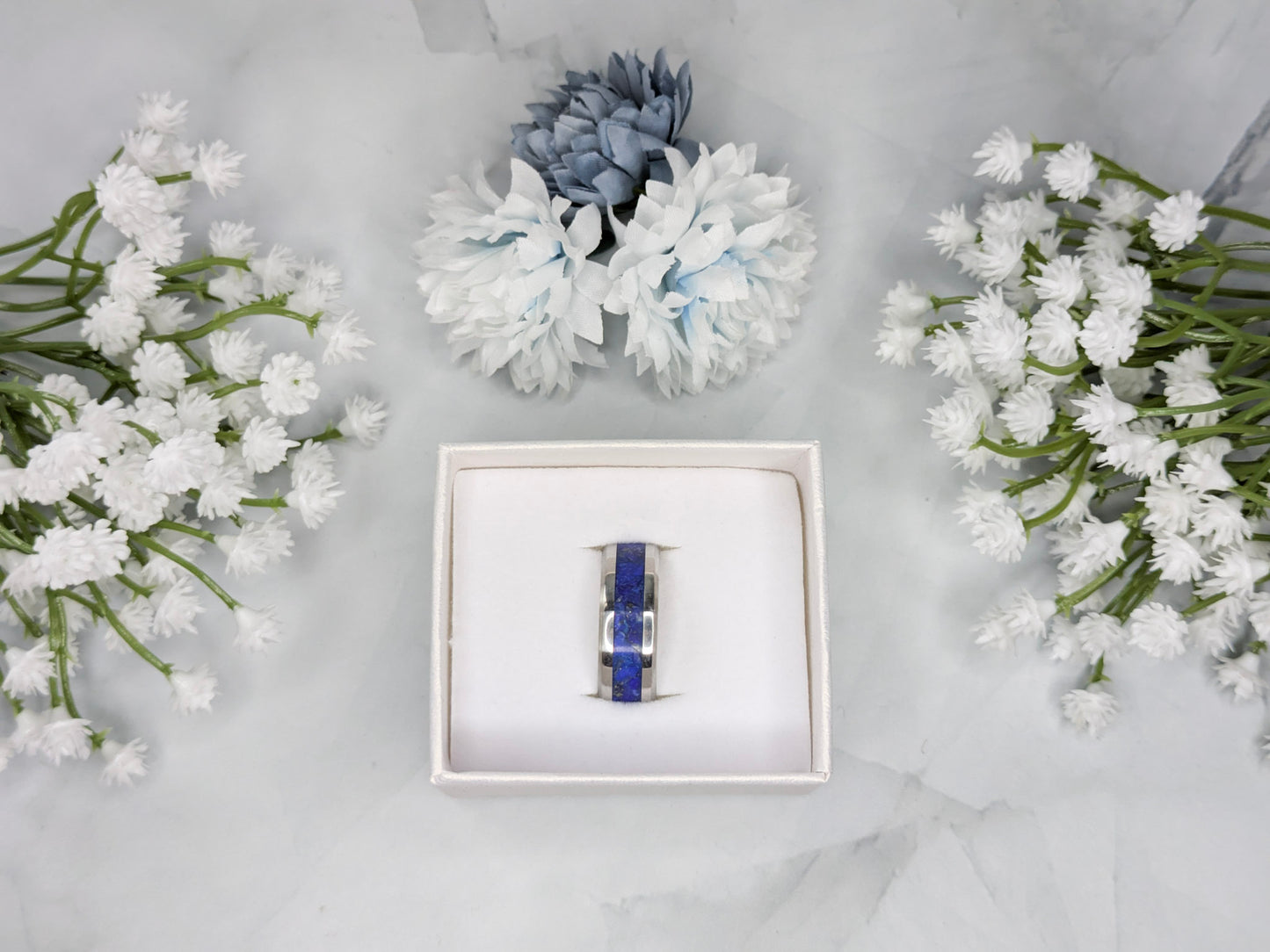 Blue lapis lazuli with flakes of gold pyrite in a stainless steel ring inlay channel. The ring is in a white jewelry box surrounded by blue and white flowers.