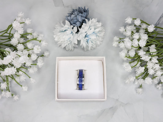 Blue lapis lazuli with flakes of gold pyrite in a stainless steel ring inlay channel. The ring is in a white jewelry box surrounded by blue and white flowers.