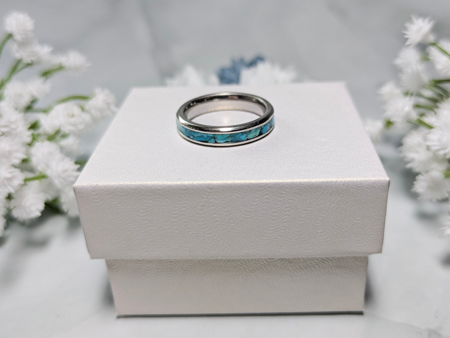 Raw natural blue Kingman turquoise in a stainless steel inlay ring channel. The ring is on top of a white jewelry box surrounded by blue and white flowers.