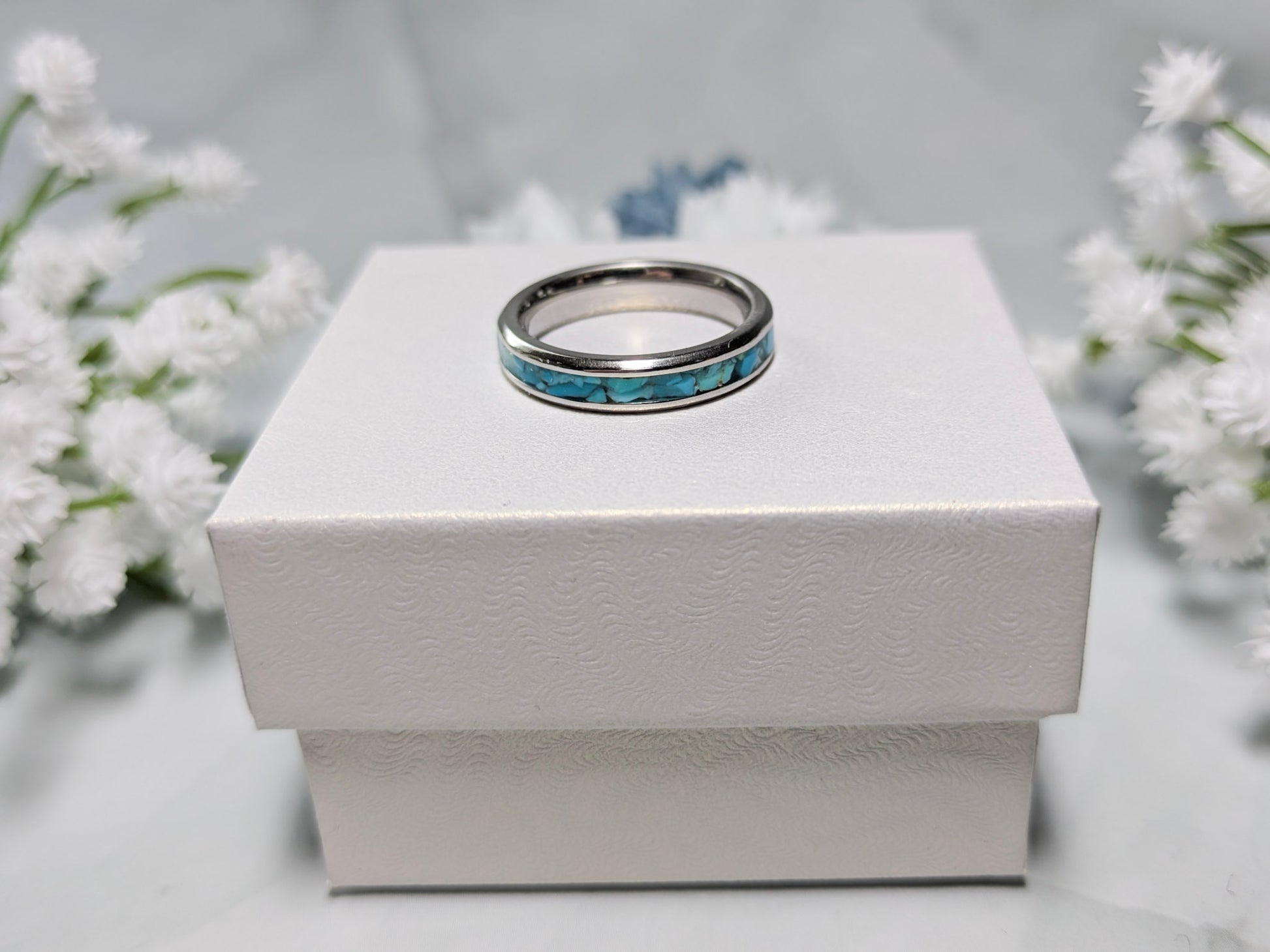 Raw natural blue Kingman turquoise in a stainless steel inlay ring channel. The ring is on top of a white jewelry box surrounded by blue and white flowers.