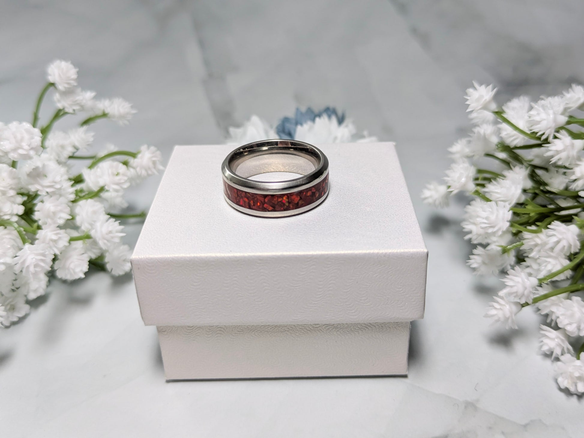 Vibrant red fire opal in a stainless steel inlay channel ring. The ring is on top of a white jewelry box surrounded by blue and white flowers.