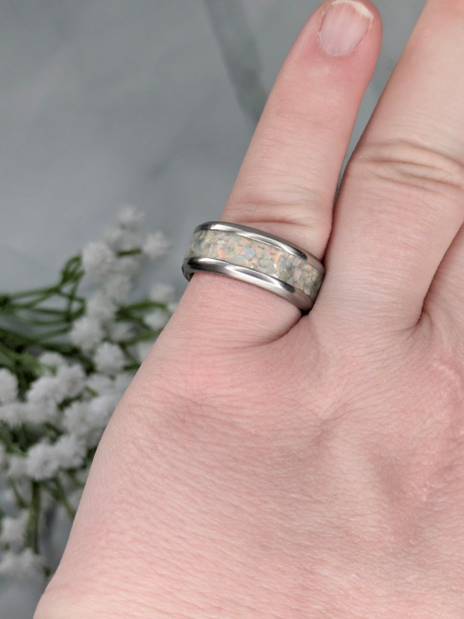 Rainbow-hued white fire and ice opal in a stainless steel inlay channel ring. The ring is on a pinky finger with white flowers in the background.
