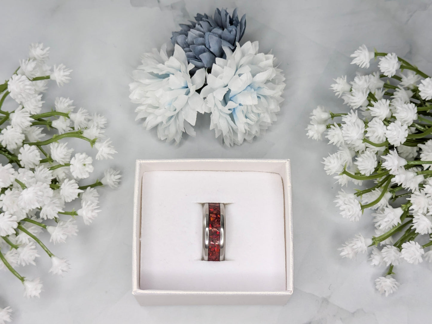 Vibrant red fire opal in a stainless steel inlay channel ring. The ring is in a white jewelry box surrounded by blue and white flowers.