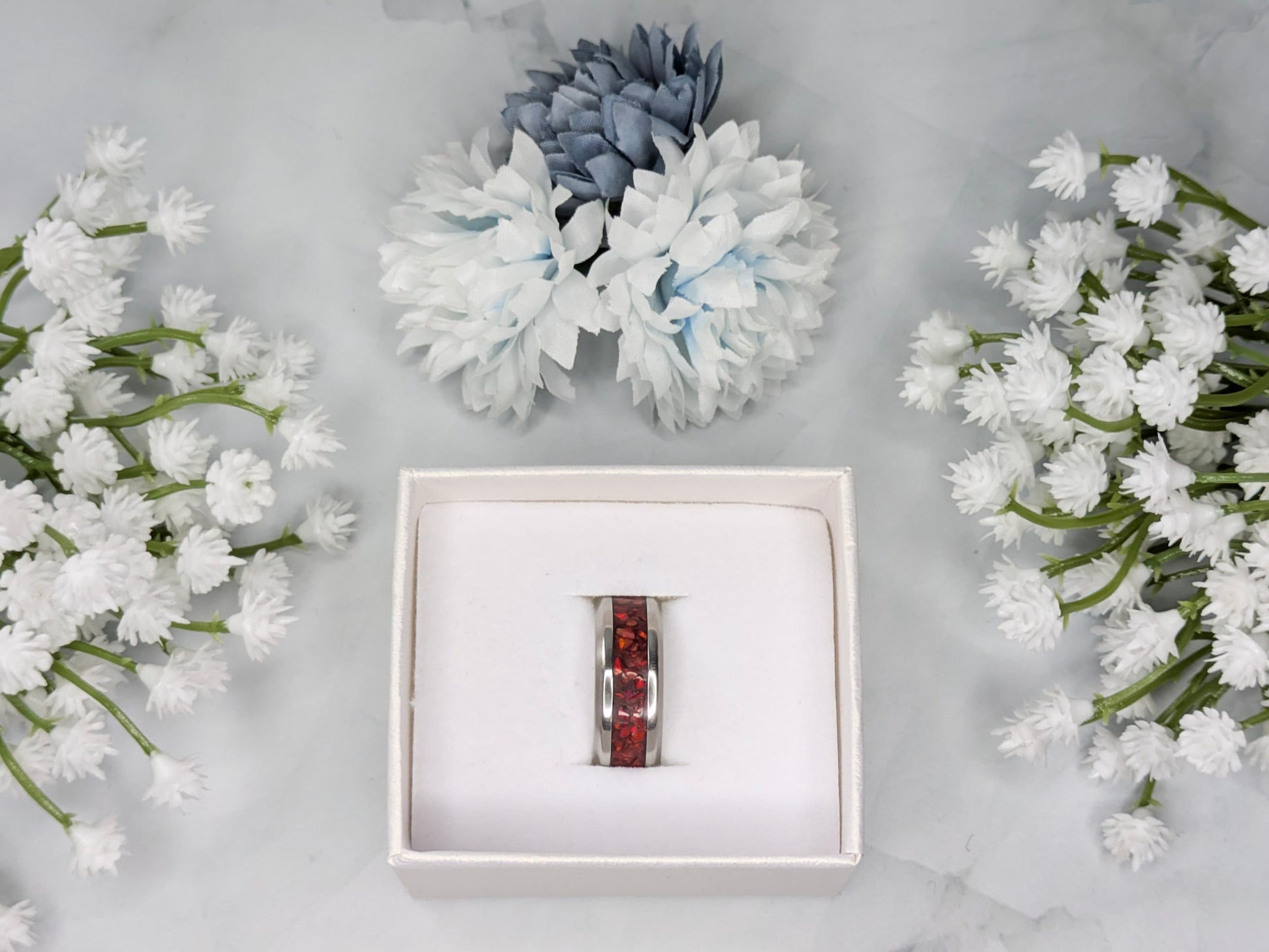 Vibrant red fire opal in a stainless steel inlay channel ring. The ring is in a white jewelry box surrounded by blue and white flowers.