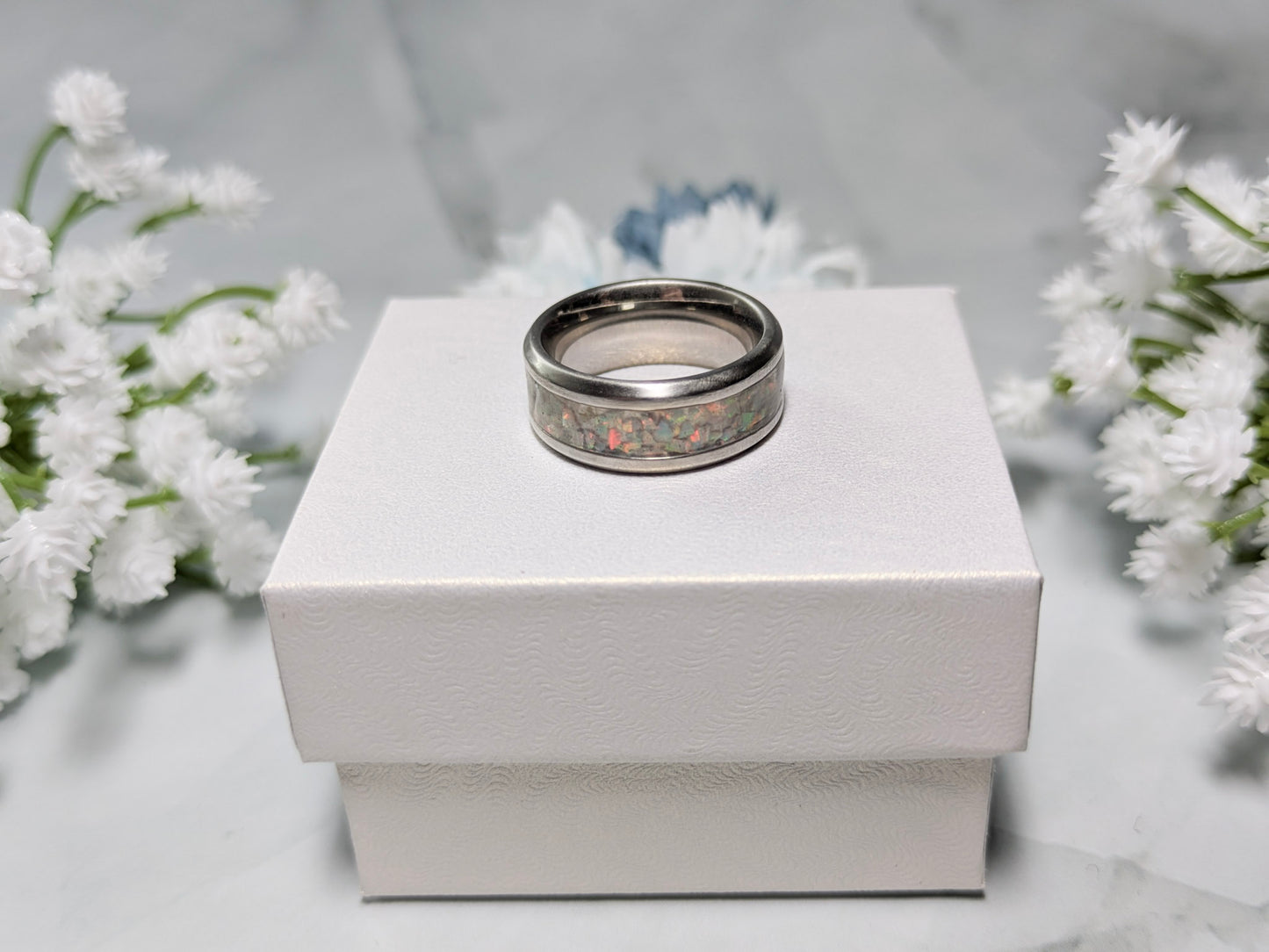 Rainbow-hued white fire and ice opal in a stainless steel inlay channel ring. The ring is on top of a white jewelry box surrounded by blue and white flowers.