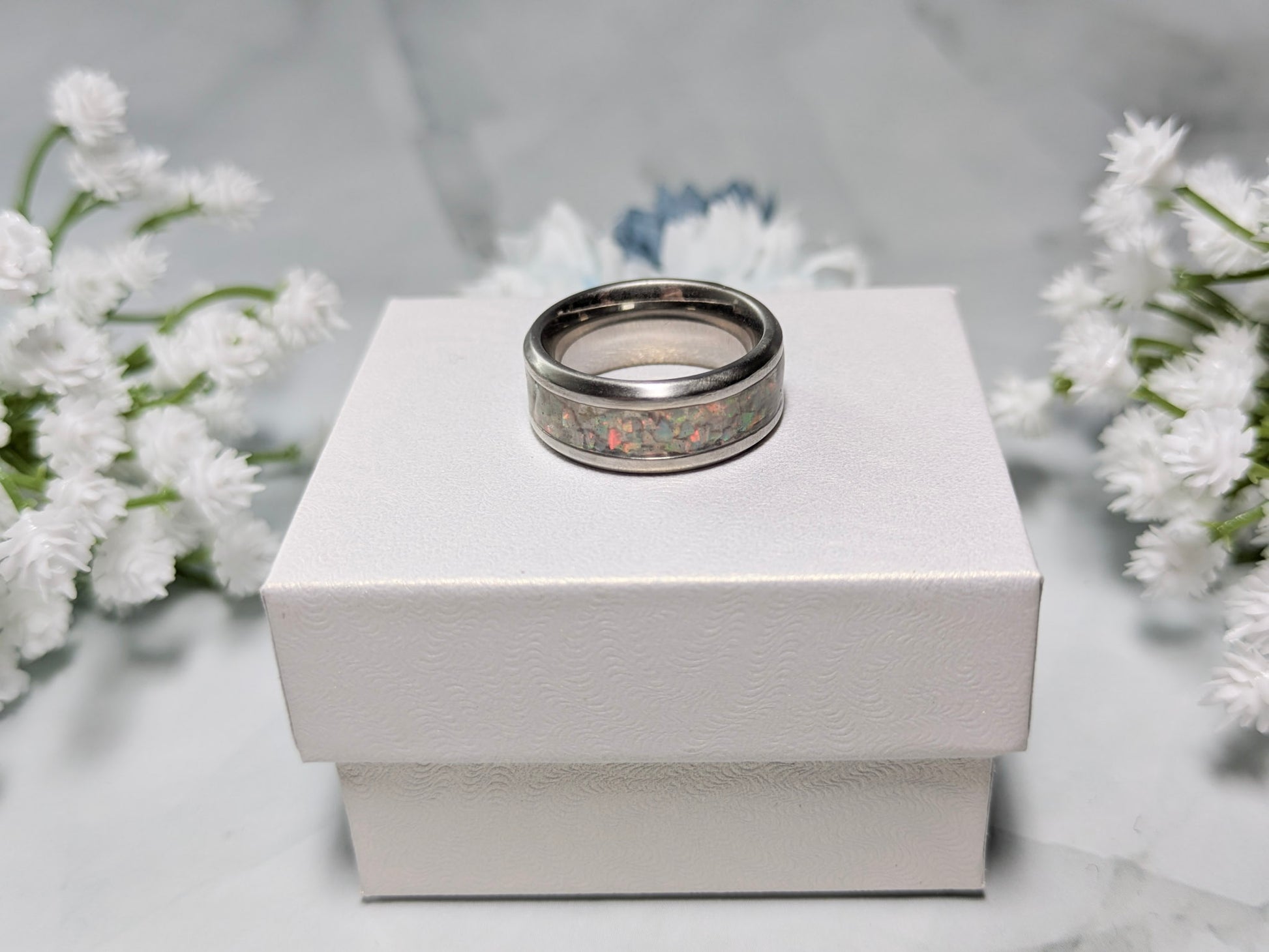 Rainbow-hued white fire and ice opal in a stainless steel inlay channel ring. The ring is on top of a white jewelry box surrounded by blue and white flowers.