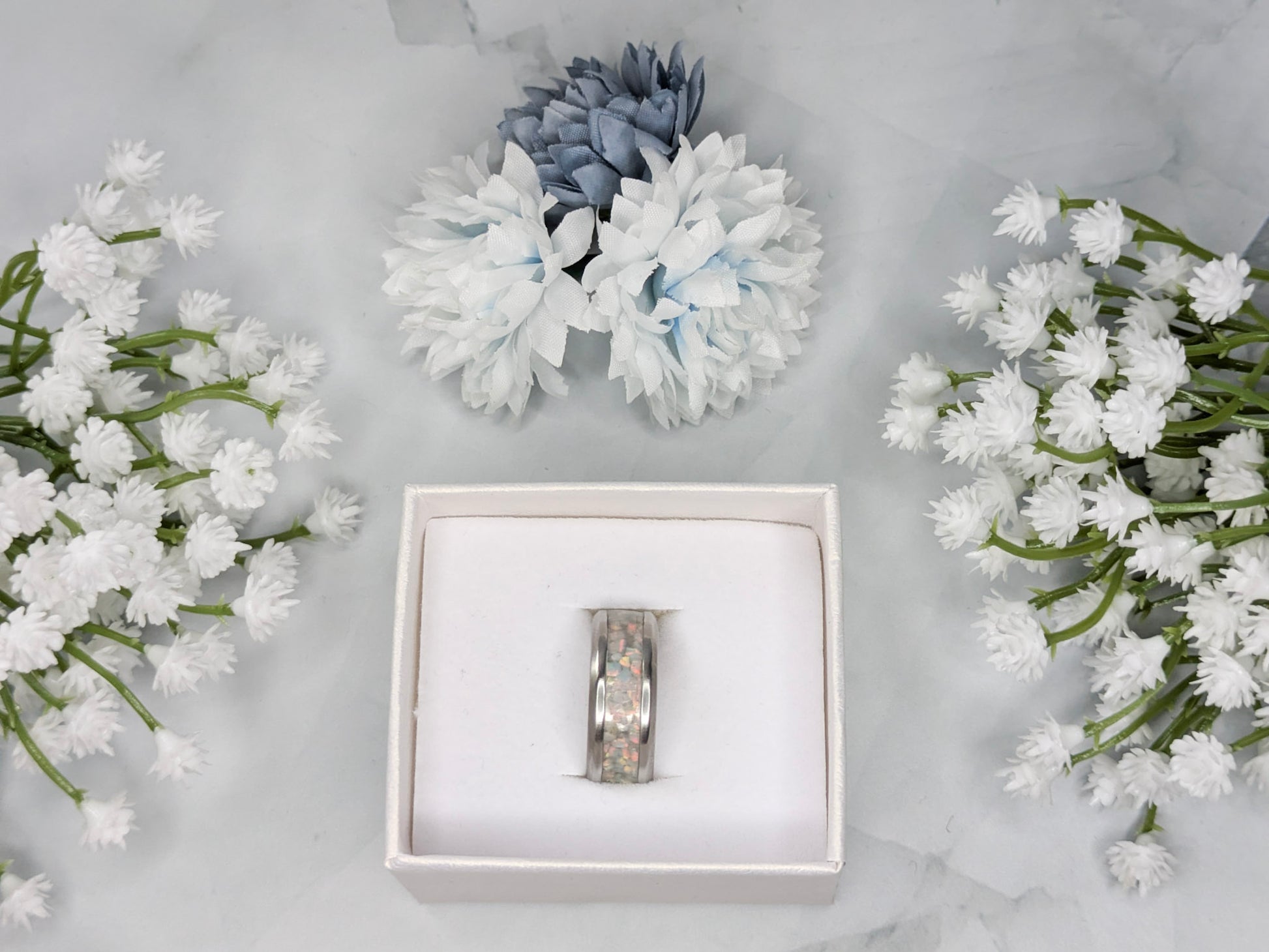 Rainbow-hued white fire and ice opal in a stainless steel inlay channel ring. The ring is in a white jewelry box surrounded by blue and white flowers.