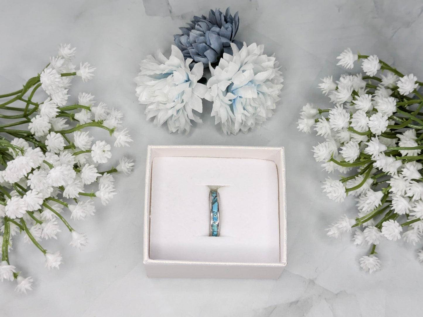Raw natural blue Kingman turquoise in a stainless steel inlay ring channel. The ring is in a white jewelry box surrounded by blue and white flowers.