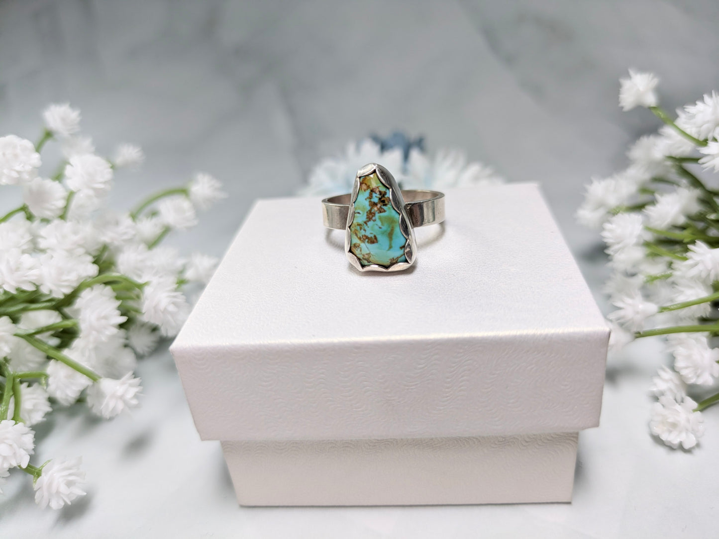Sterling silver ring with a triangular turquoise gemstone. The turquoise is a rich sky blue and leaf green with specks of brown. The ring is on top of a white jewelry box surrounded by white and blue flowers.