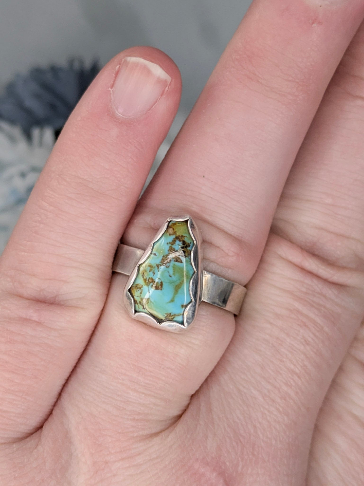 Sterling silver ring with a triangular turquoise gemstone. The turquoise is a rich sky blue and leaf green with specks of brown. The ring is on a finger with flowers in the background.