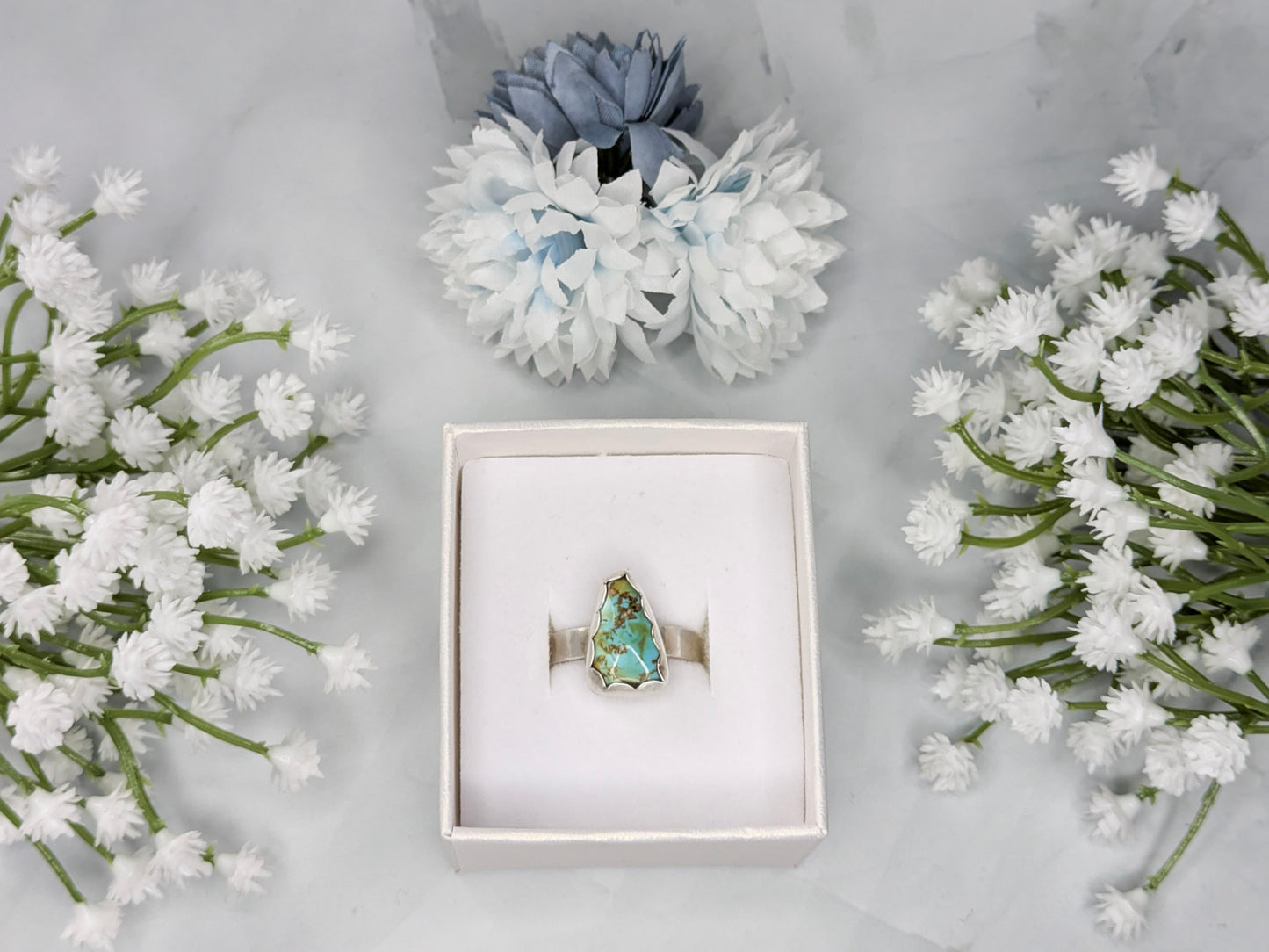 Sterling silver ring with a triangular turquoise gemstone. The turquoise is a rich sky blue and leaf green with specks of brown. The ring is in a white jewelry box surrounded by white and blue flowers.