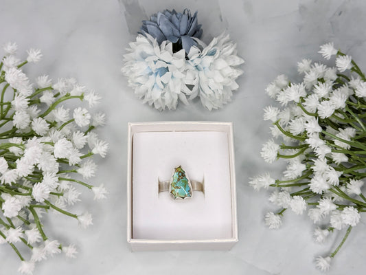 Sterling silver ring with a triangular turquoise gemstone. The turquoise is a rich sky blue and leaf green with specks of brown. The ring is in a white jewelry box surrounded by white and blue flowers.