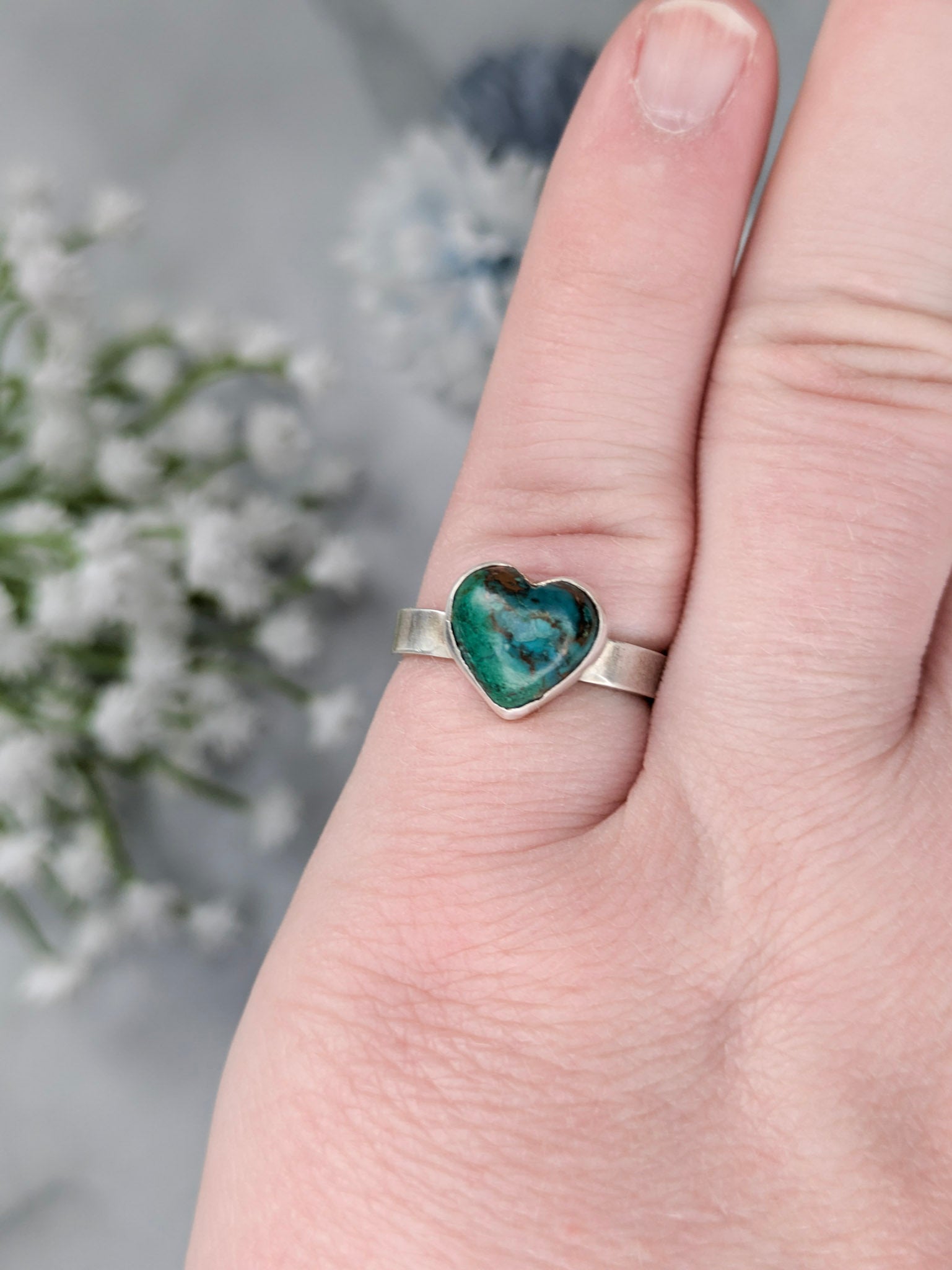 Sterling silver ring with a heart-shaped blue-green chrysocolla gemstone. The ring is worn on a pinky finger with flowers in the background.