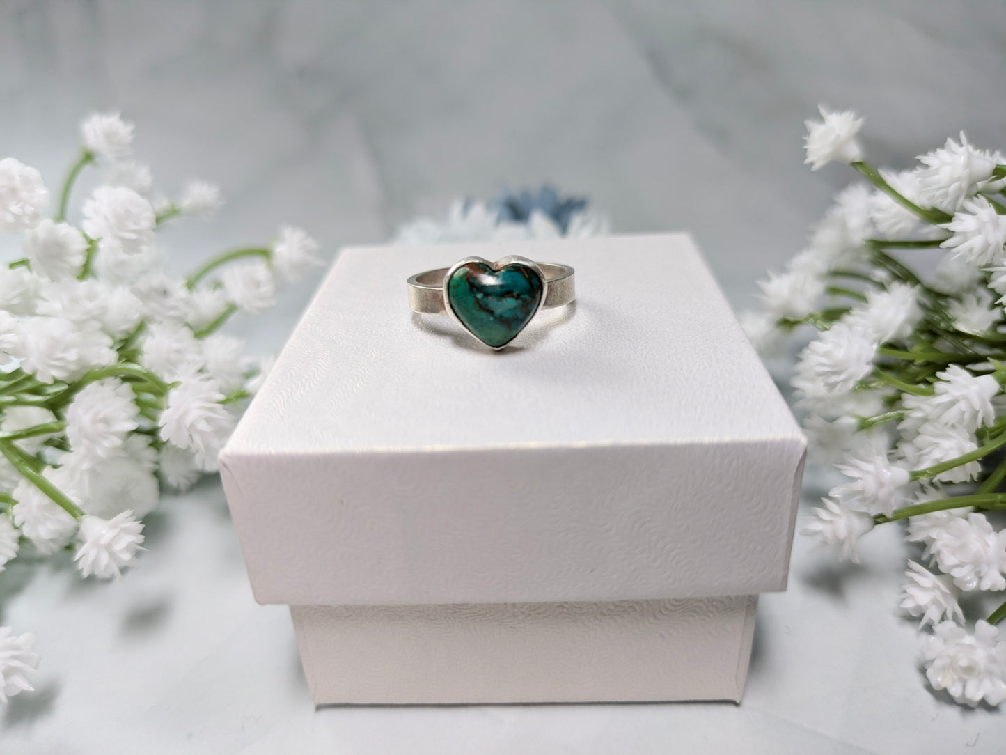 Sterling silver ring with a heart-shaped blue-green chrysocolla gemstone. The ring is on top of a white jewelry box surrounded by blue and white flowers.