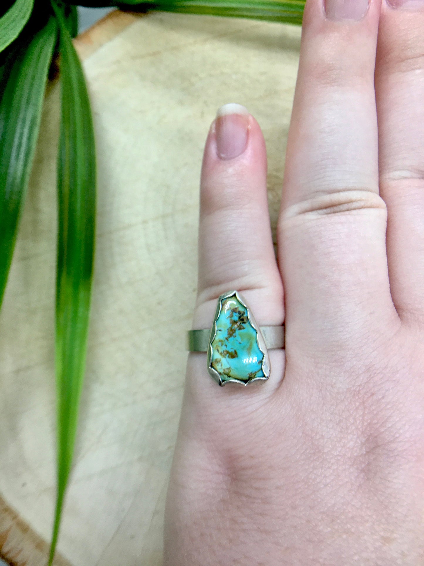 Sterling silver ring with a triangular turquoise gemstone. The turquoise is a rich sky blue and leaf green with specks of brown. The ring is on a finger with wood and green plants in the background.