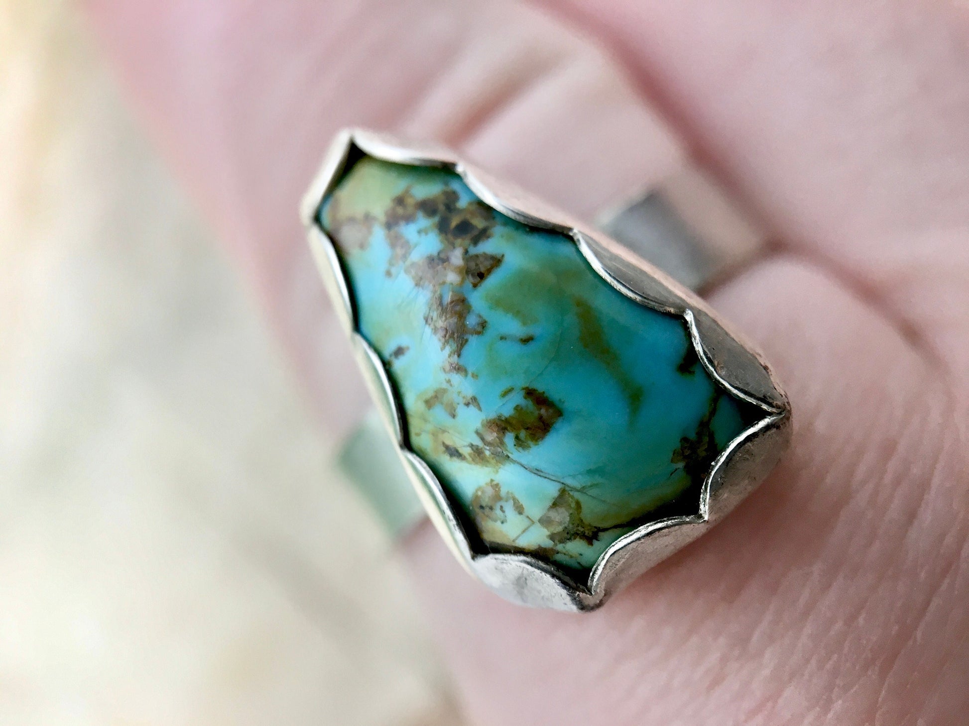 Close up of a sterling silver ring with a triangular turquoise gemstone. The turquoise is a rich sky blue and leaf green with specks of brown. The ring is on a finger.