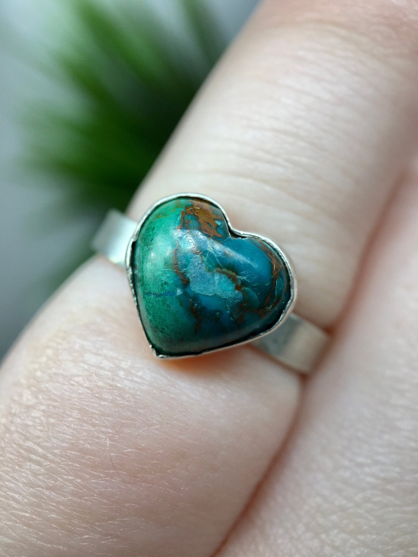 Sterling silver ring with a heart-shaped blue-green chrysocolla gemstone. The ring is on a pinky finger with green plants in the background.